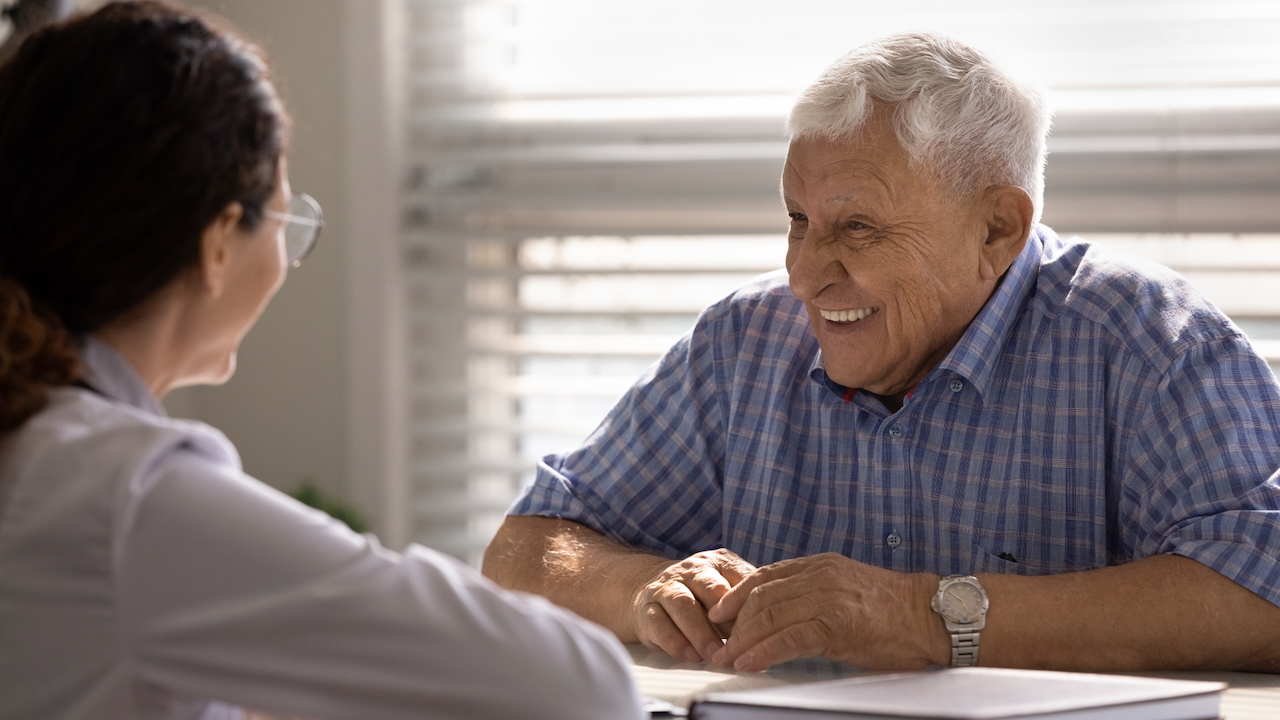 smiling mature 80s male patient talk with caring female nurse in hospital. Happy senior man patient have good quality consultation in Savya clinic.